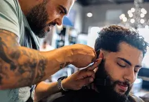 A barber applies an electric razor to the hair behind the ear of a customer. 
